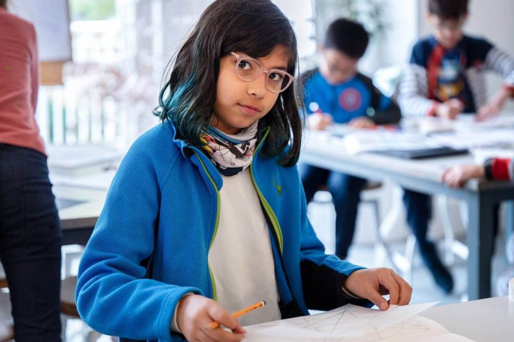 A young female student with a blue jacket and glasses looking at the camera whilst holding her drawing and a pencil